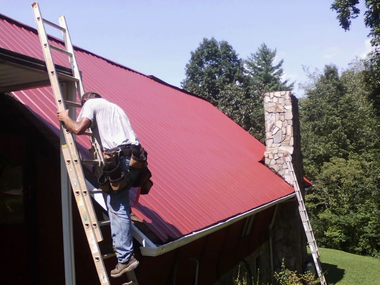 Roofer on a ladder beside a red metal roof and stone chimney — Soffit & Fascia Repair by Roofing Brothers in Mantua